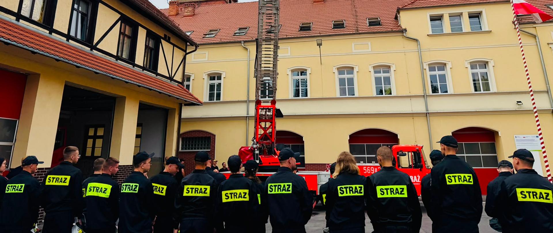 Na zdjęciu widać strażaków stojących na placu przed komendą. Strażacy uczą się musztry. Strażacy są ubrani w mundury koszarowe koloru czarnego i służbowe koloru musztardowego, strażacy stoją w dwu szeregu. Strażacy stoją przed drabiną. W tle widać budynek komendy. 