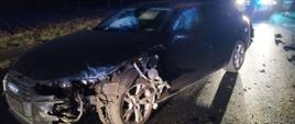 Zderzenie dwóch aut. Droga wojewódzka nr 309 w okolicach Żylic. Na jezdni stoi rozbite audi. Pojazd ma uszkodzoną swoją przednią - lewą stronę. Auto jest koloru ciemnego. Na jezdni znajdują się oderwane od samochodu elementy. W tle widoczny strażak i policjant.