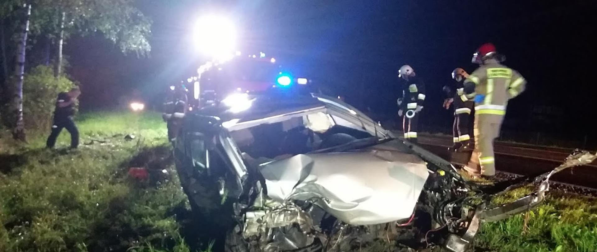 Noc. Zniszczony w wyniku wypadku samochód znajduje się na trawie na poboczy drogi. Wokół niego funkcjonariusze oraz strażacy prowadzą czynności. W tle znajduje się samochód strażacki, który oświetla teren zdarzenia.