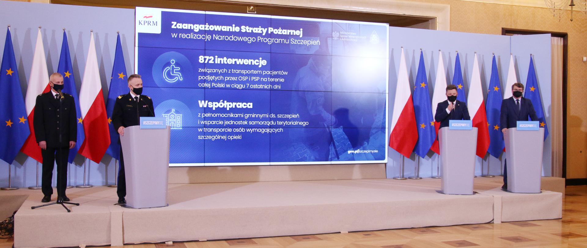 Komendant Główny PSP wraz z przedstawicielami rządu przy mównicach na tle banneru Narodowego Programu Szczepień.