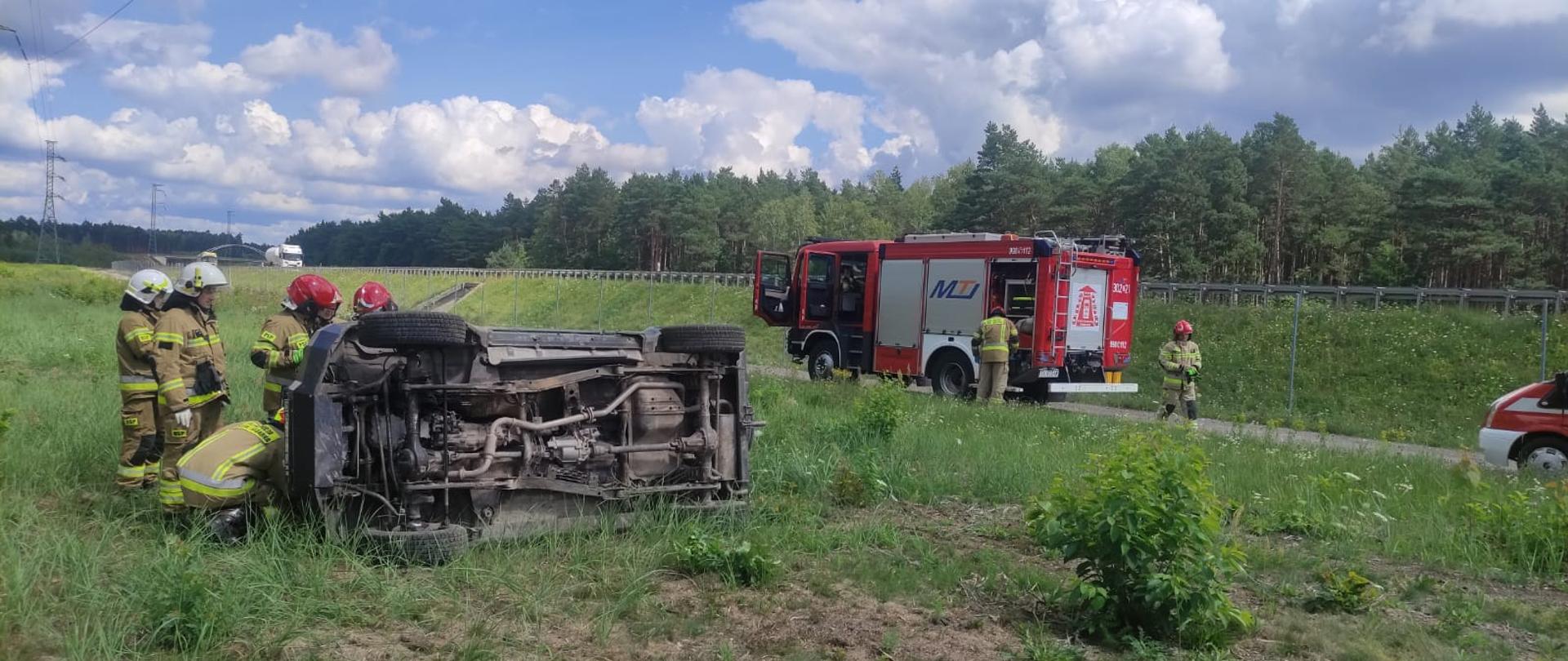 Zdjęcie przedstawia miejsce wypadku. W centralnym punkcie obrazu leży na boku samochód osobowy przy którym stoi grupa strażaków, w głębi widać samochód pożarniczy i dwóch ratowników oraz fragment drugiego pojazdu strażackiego.