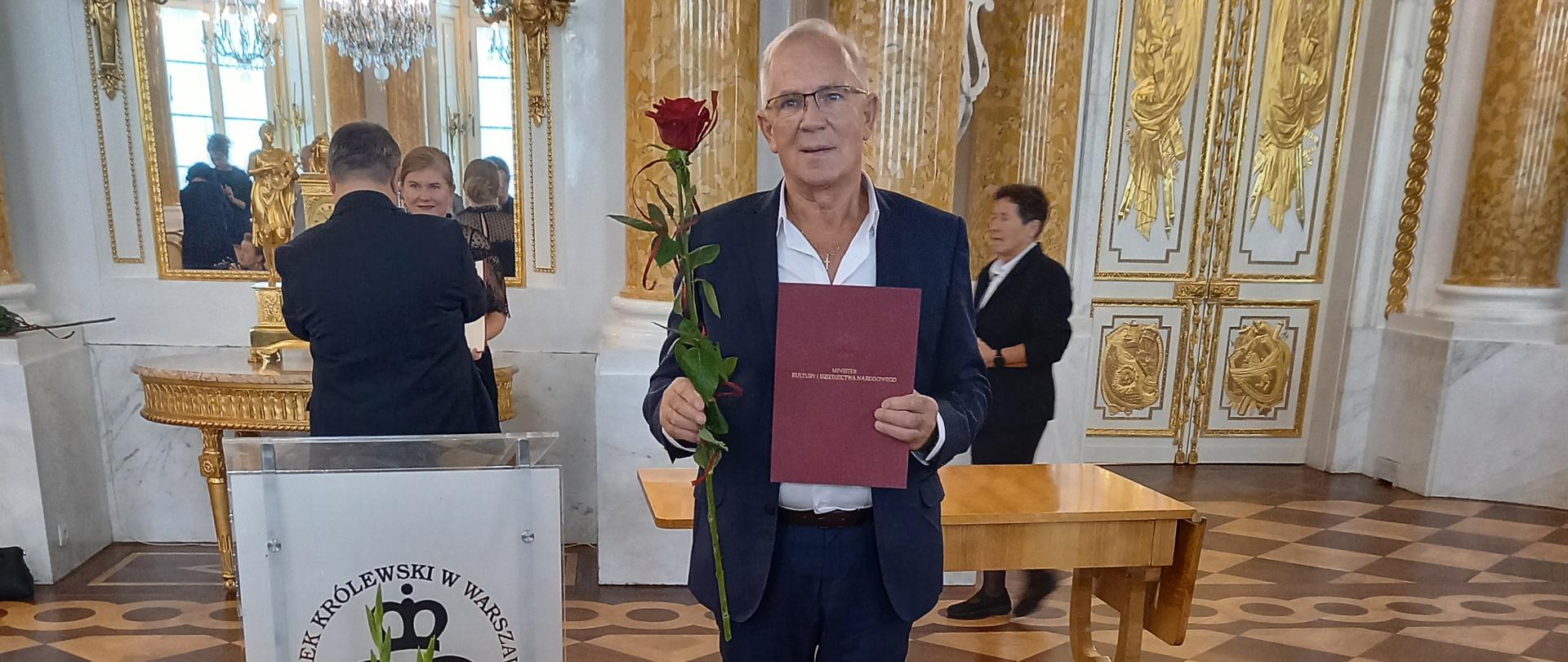 Nauczyciel stoi przy mównicy ozdobionej wiązanką biało – czerwonych kwiatów. W rękach trzyma dokument i czerwoną różę. W tle znajdują się osoby biorące udział w uroczystości. Sala Zamku Królewskiego utrzymana w brązowo – złoto – białym kolorze.