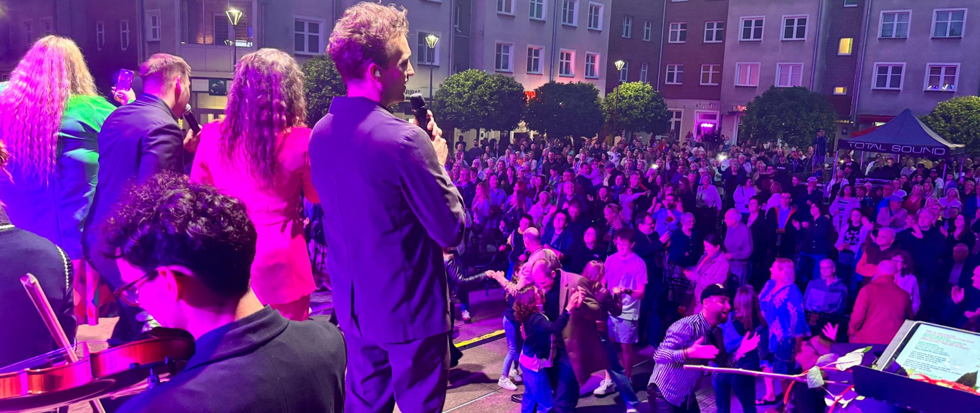 Liczna widownia stojąca oraz tańcząca pod sceną. Na scenie przodem do publiczności stoją i śpiewają wokaliście. Zewsząd pada różowe światło sceniczne. W tle wysokie budynki mieszkalne.