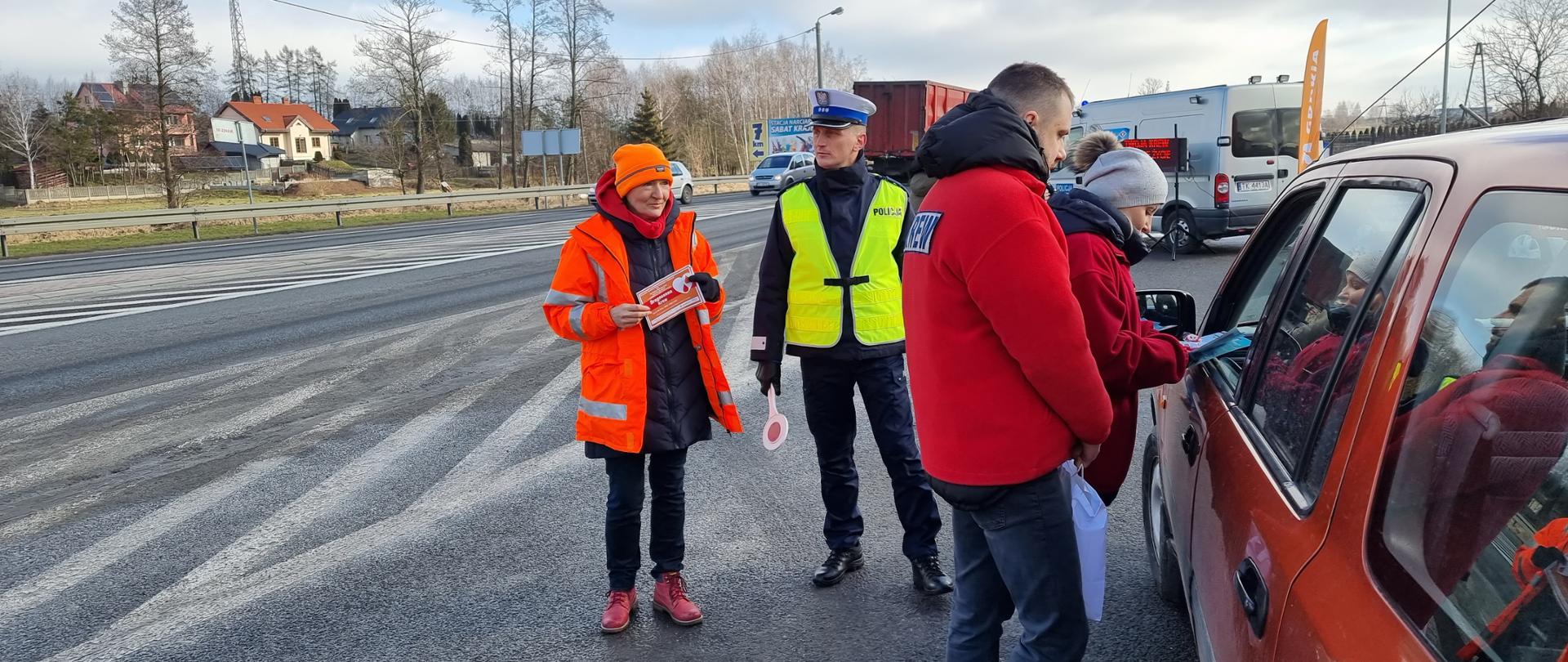 Akcja "Drogocenna krew" - policjant, drogowiec, pracownicy centrum krwiodawstwa rozmawiają z kierowcą w samochodzie, w tle droga 