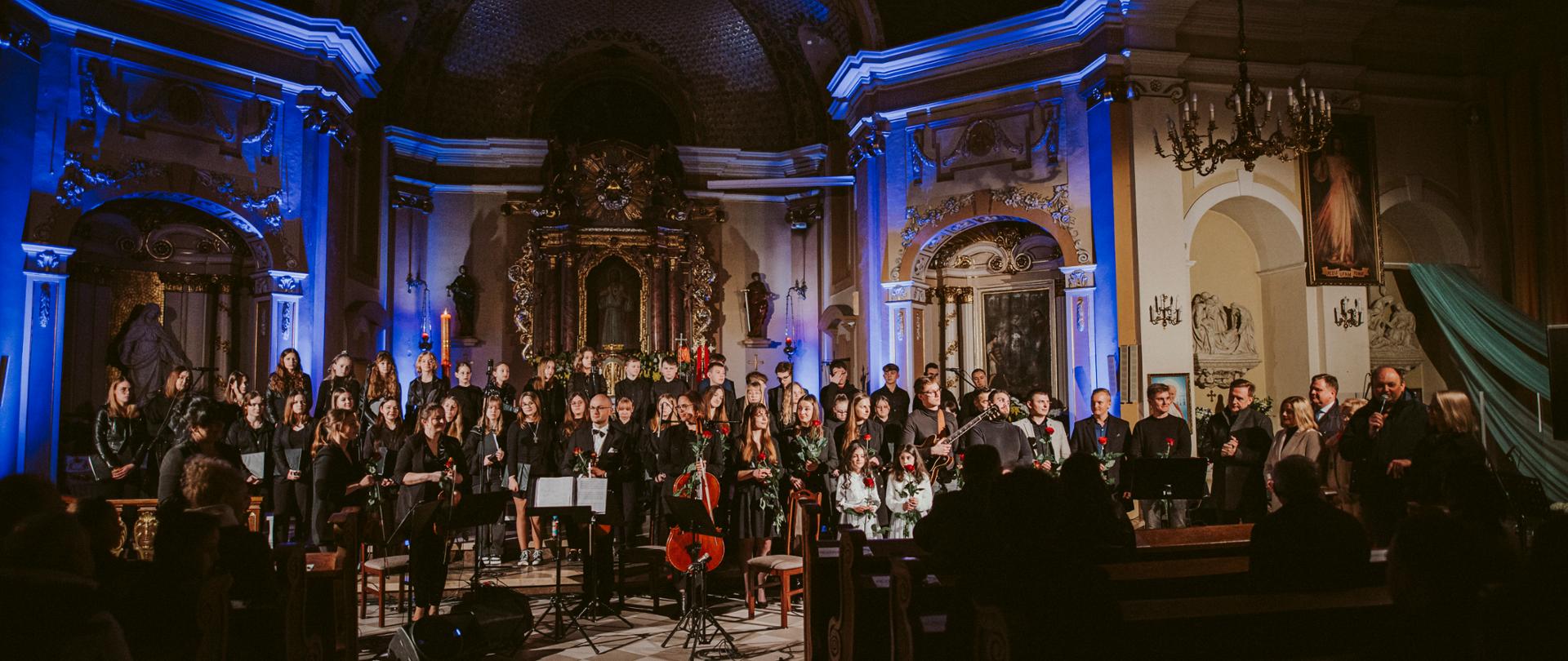 Wszyscy wykonawcy Koncertu Wielkanocnego w kościele św. Marcina w Kępnie na tle ołtarza. Z tyłu w kilku rzędach stoją chórzyści, z przodu od lewej stoi kwartet smyczkowy, dalej soliści i Ekipa Dobrego Dźwięku, natomiast od prawej Starosta Kępiński wraz z Zarządem Powiatu. Tło dopełnia niebieskie oświetlenie. 