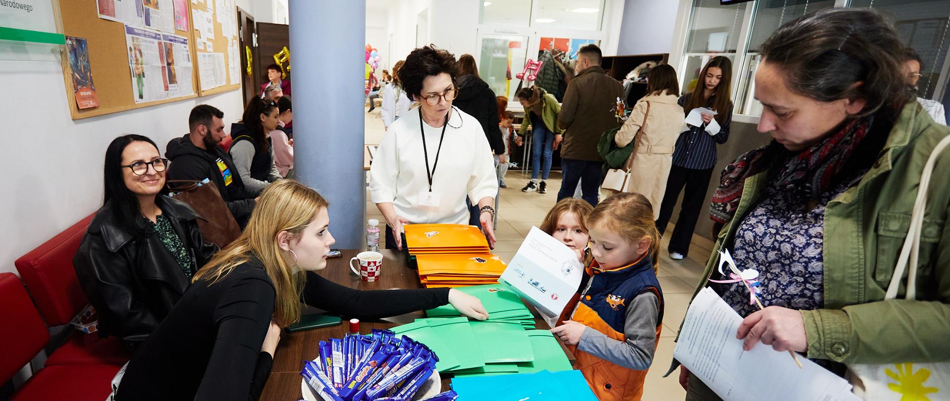Dzieci i dorośli stoją przy biurku, na którym leżą teczki z upominkami na Dzień Otwarty Szkoły