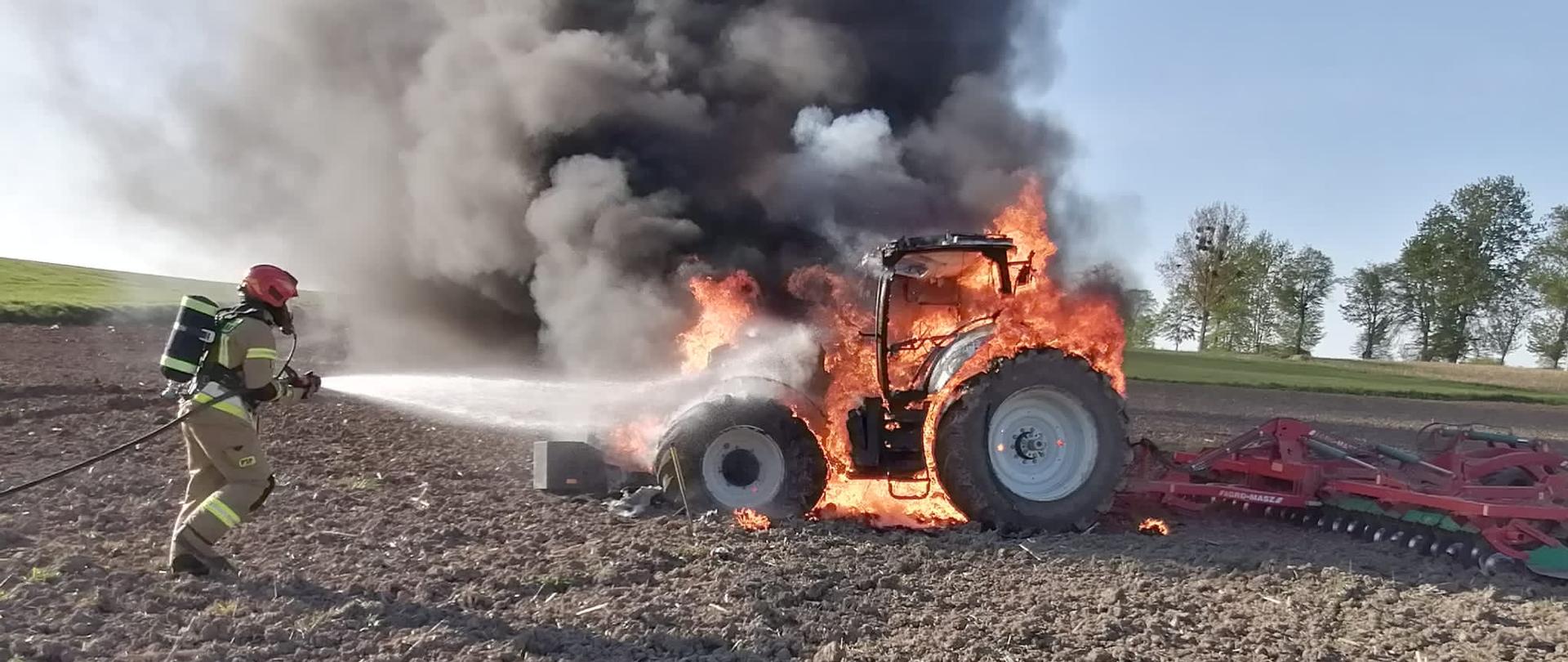 Pożar ciągnika rolniczego na uprawianym polu. Ciągnik cały w ogniu, dużo dymu. Strażak podchodzi z linia gaśniczą.