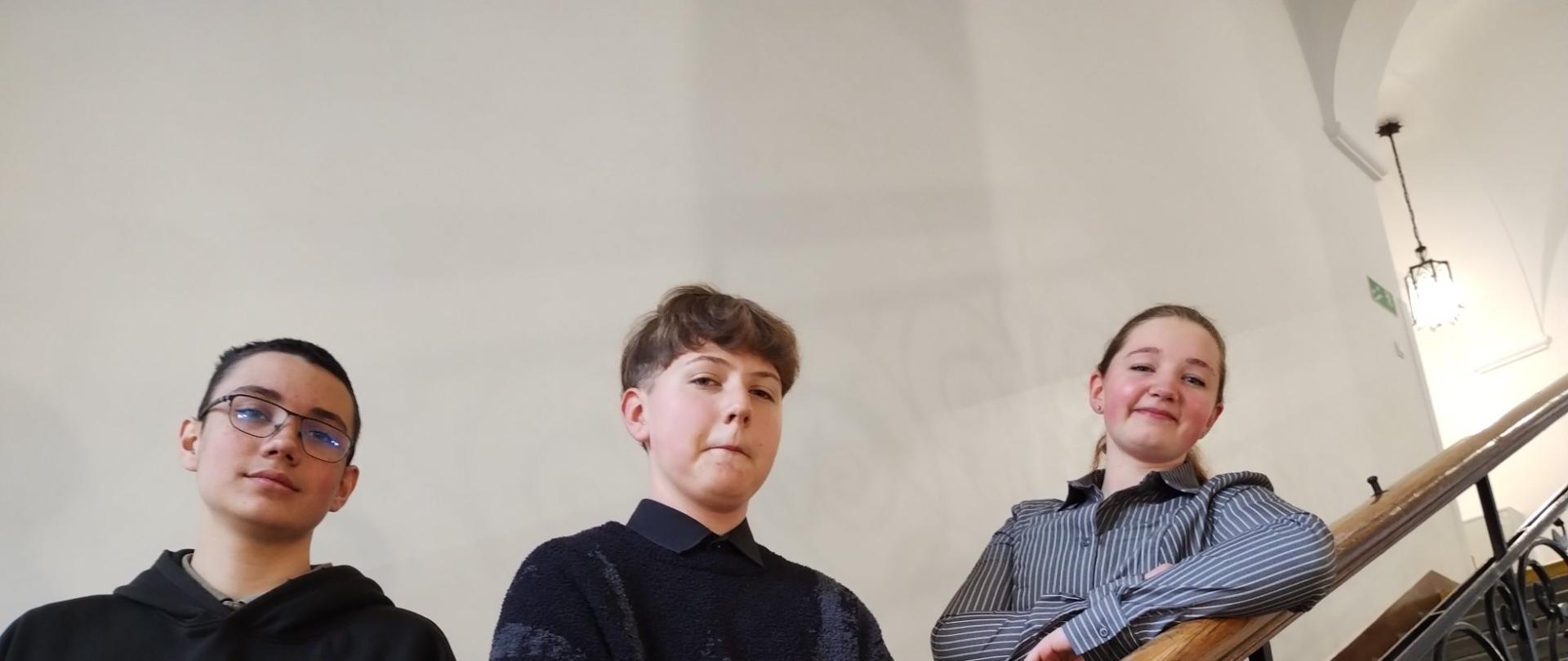 Trzy osoby stoją na klatce schodowej; widoczne drewniane poręcze i metalowe balustrady.