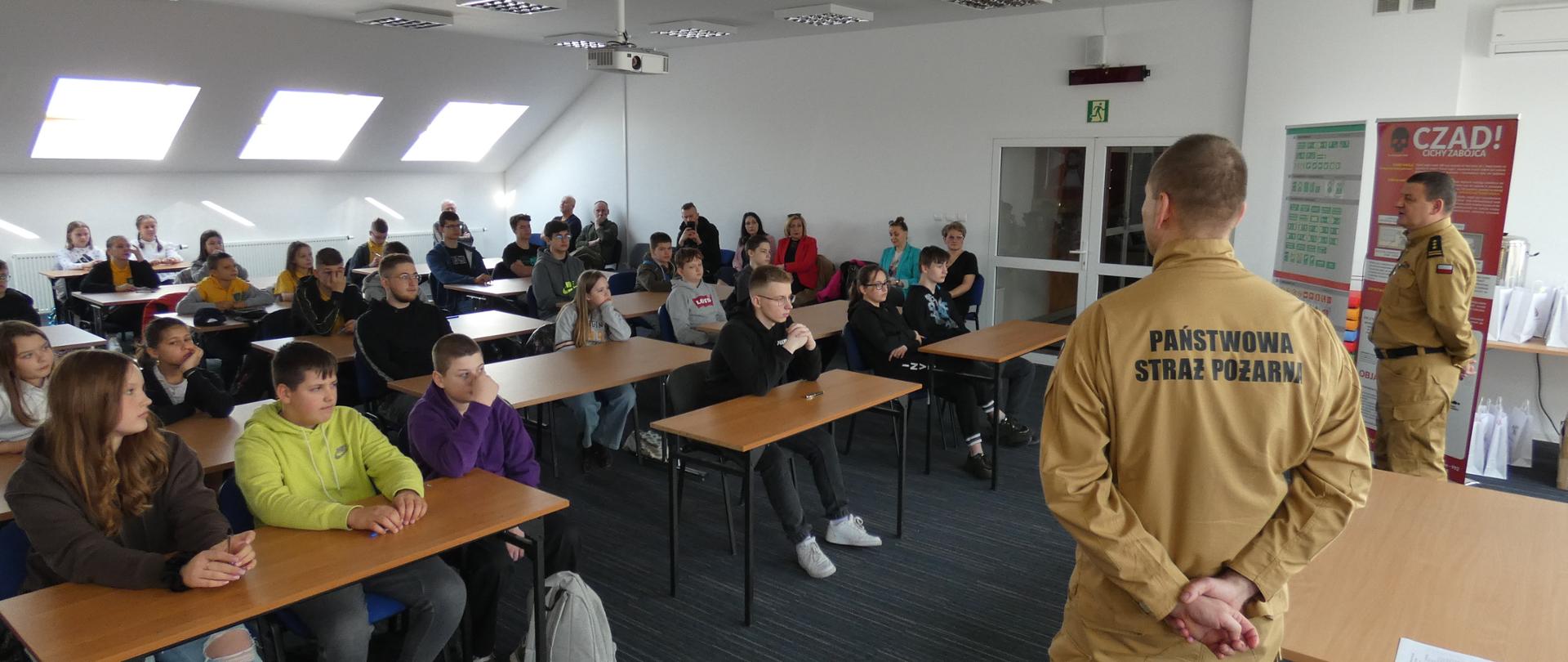 Młodzież siedząca w ławkach wysłuchuje oficjalnego rozpoczęcia konkursu. Przemawia Zastępca Komendanta Powiatowego PSP w Ełku st. bryg. Paweł Litwinowicz.