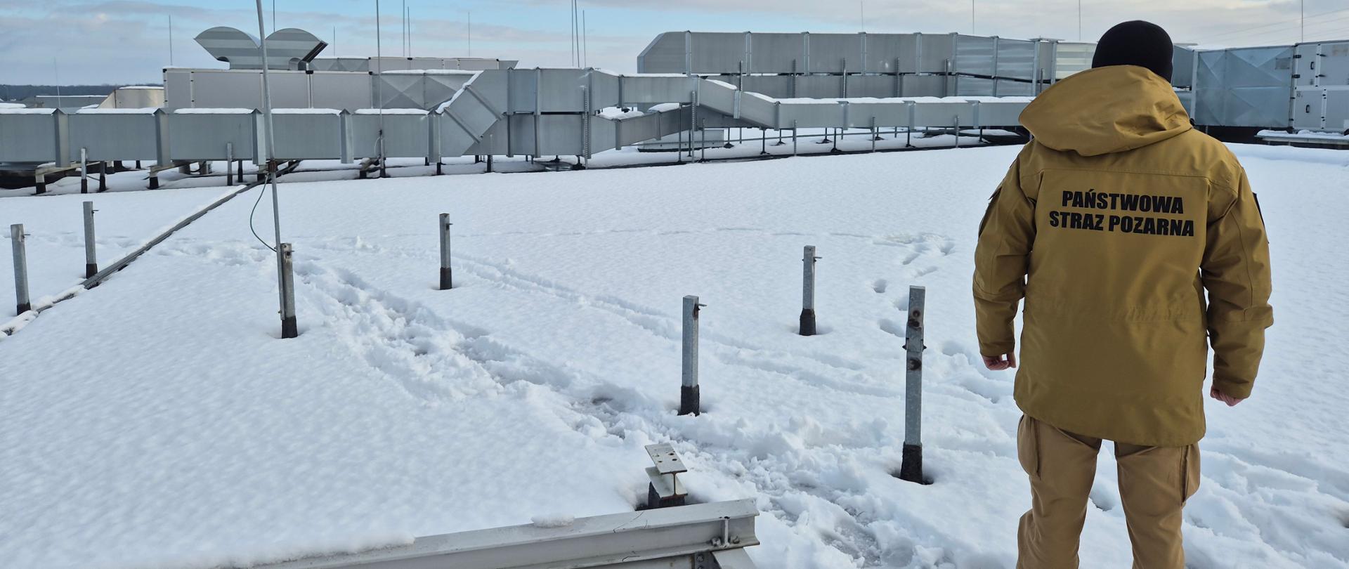 Dach budynku z pokrywą śnieżną. Strażak stoi tyłem na dachu. Z daleka widać przewody wentylacji mechanicznej. 