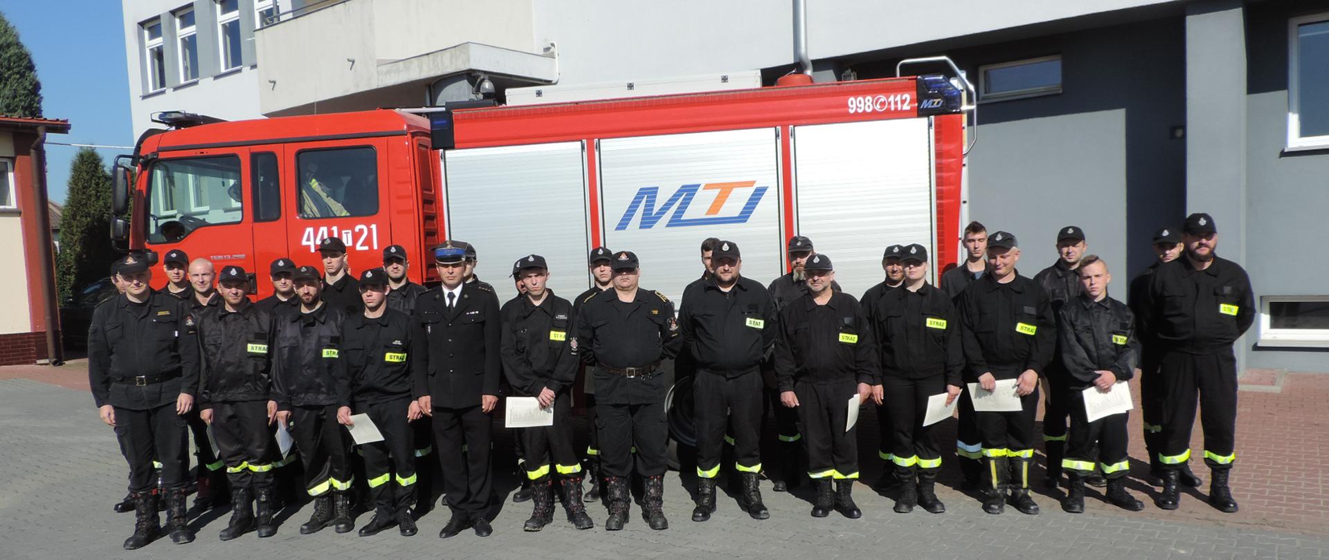 Zdjęcie przedstawiające komendanta powiatowego wraz z druhami OSP uczestniczącymi w kursie podstawowym ustawionych na zbiórce przy samochodzie ciężkim gaśniczym na placu KP PSP w Opatowie