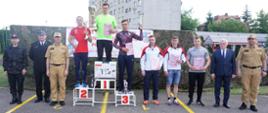 Na zdjęciu są zawodnicy stojący na podium oraz strażacy wręczający puchary 