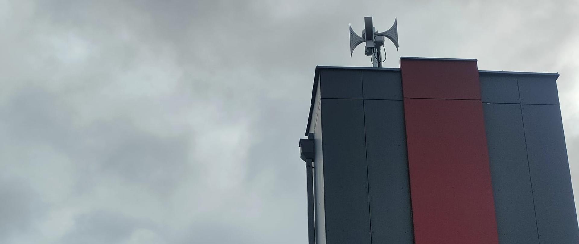 Budynek Komendy Powiatowej Państwowej Straży Pożarnej, na wieży widoczne tuby rozgłoszeniowe systemu ostrzegania i alarmowania, dach budynku pokryty blachą, elewacja wykonana z płyt koloru szarego i bordowego, na elewacji wieży zamontowany herb strażacki w kolorze czarnym 