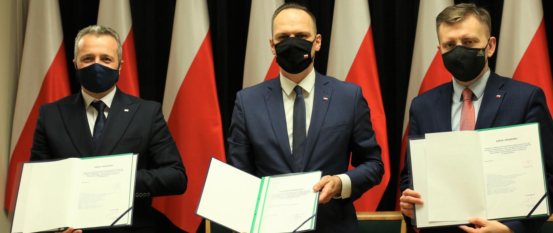 Wiceminister Rafał Weber oraz minister Łukasz Schreiber i wojewoda Mikołaj Bogdanowicz prezentują decyzję o finansowaniu ronda w Kobylnikach.