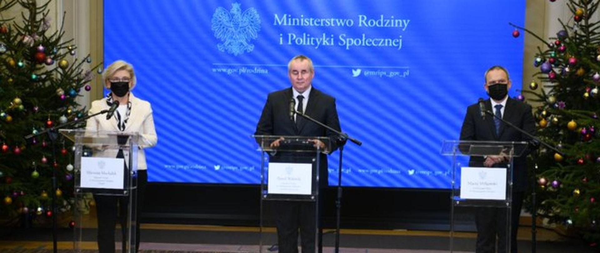 trzy osoby stoją przed mównicami po obu stronach stoją choinki, a z tyłu wyświetla się prezentacja. 