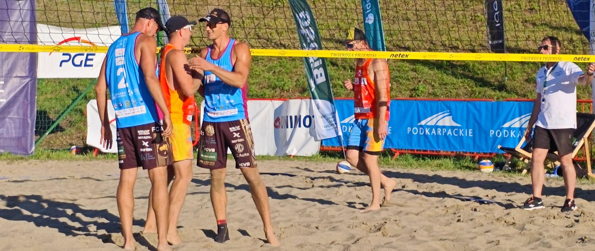 Zawodnicy obu drużyn spotykają się przy siatce po rozegranej akcji podczas turnieju siatkówki plażowej. W tle widoczne są banery partnerów wydarzenia.
