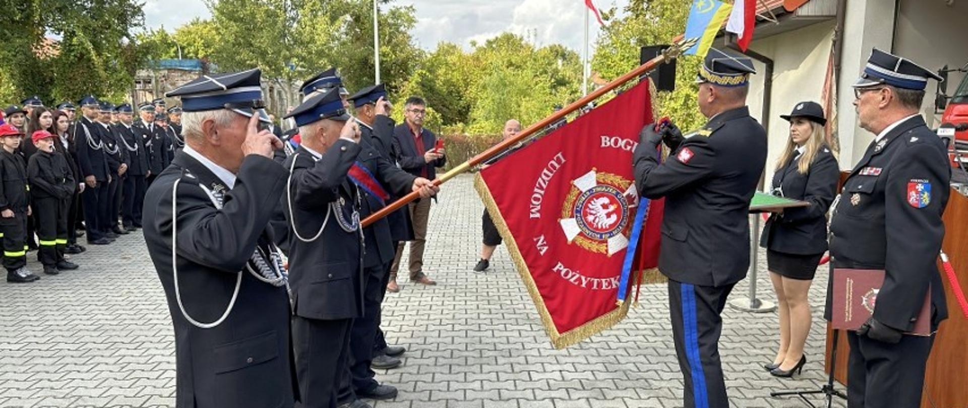 Zdjęcie zrobione na zewnątrz, podczas uroczystych obchodów jubileuszu jednostki Ochotniczej Straży Pożarnej. Generał Państwowej Straży Pożarnej odznacza medalem sztandar jednostki OSP. 