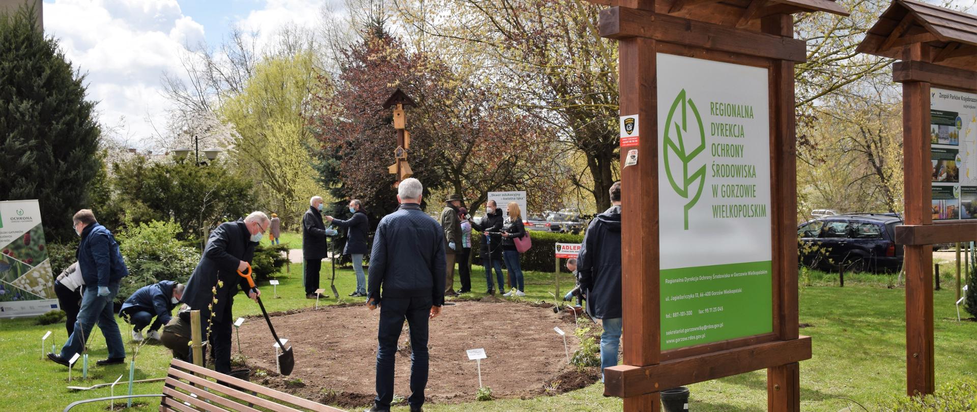 Na zdjęciu tablica z Logo RDOŚ, w oddali zgromadzeni goście spoglądają na sadzone już byliny. Zgromadzeni goście udzielają wywiadu mediom na skwerze edukacyjnym