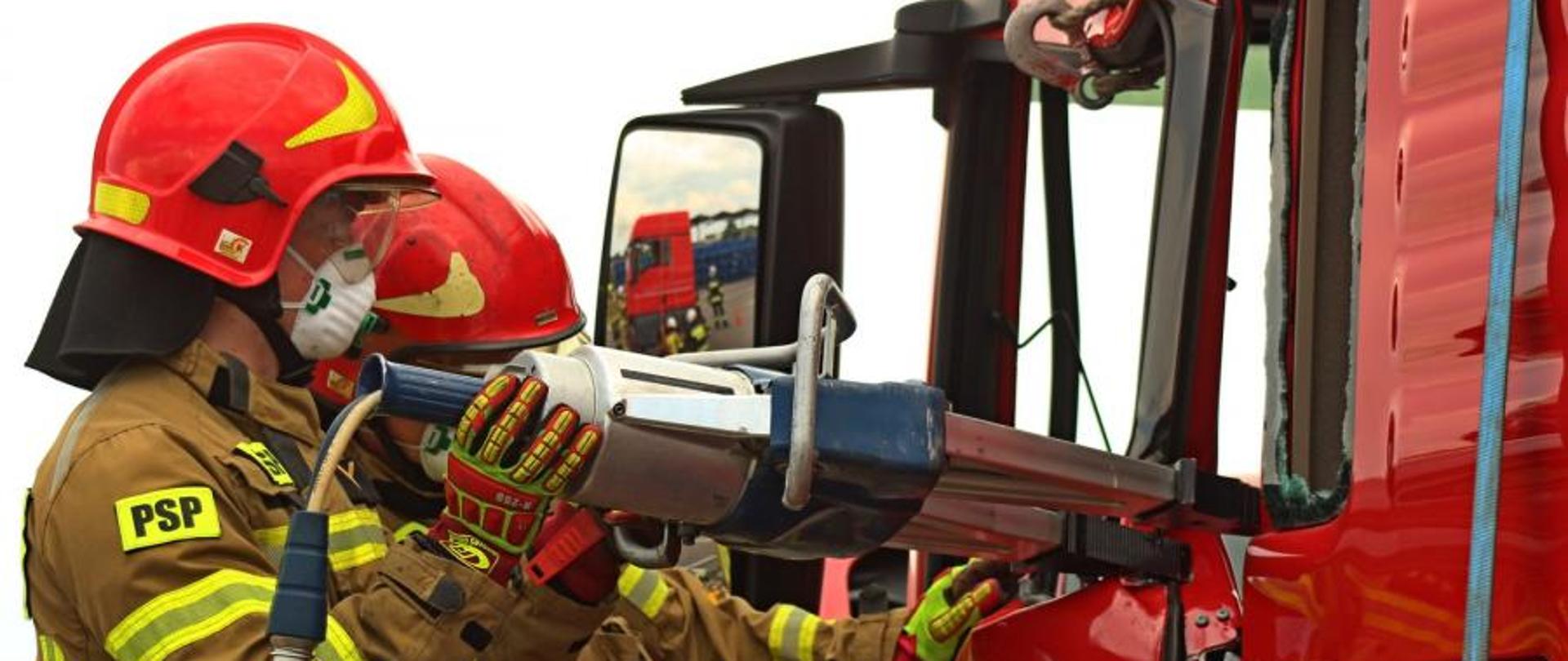 Widoczni dwaj strażacy w umundurowaniu specjalnym oraz część samochodu ciężarowego. Strażacy w rękach trzymają rozpieracz hydrauliczny, którym wyważają drzwi do samochodu ciężarowego.