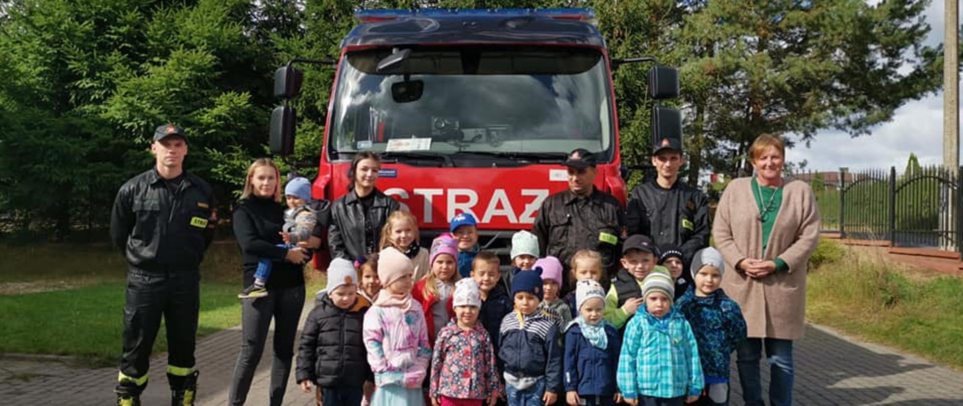 Grupa dzieci z opiekunami i trzema strażakami w ubraniach koszarowych stoją na tle wozu strażackiego.