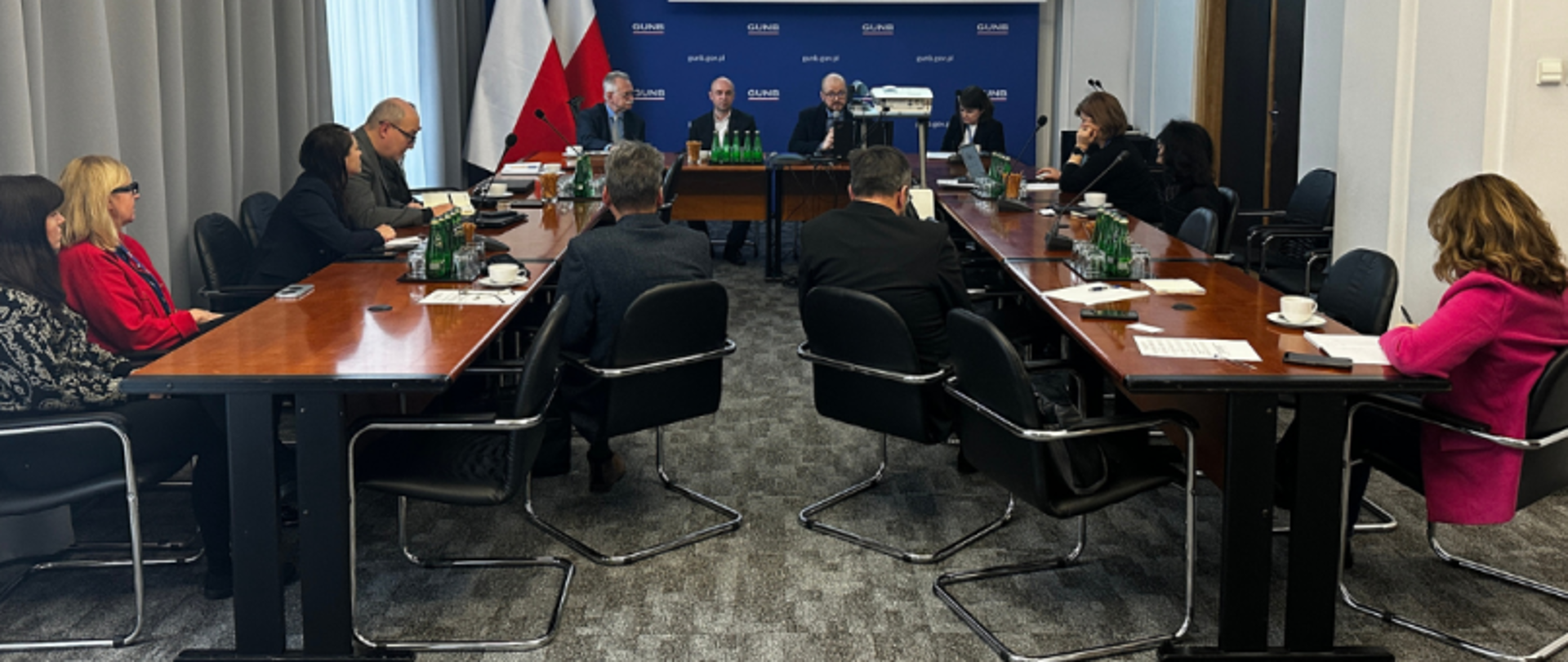 Grupa osób siedzi przy stołach w kształcie litery "U" podczas formalnego spotkania w sali konferencyjnej. Na końcu sali znajduje się stół prezydialny z trzema mężczyznami, za nimi widnieje niebieskie tło z logo instytucji oraz polska i unijna flaga. Na stołach znajdują się dokumenty, butelki wody oraz filiżanki z kawą. Pomieszczenie ma szare zasłony i wykładzinę dywanową.