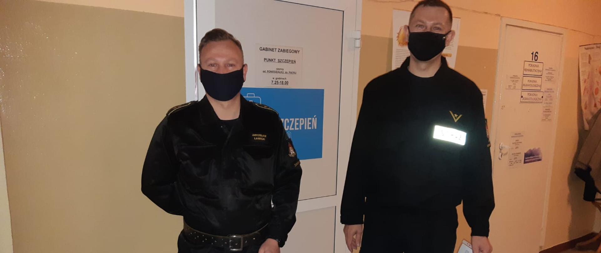 Dwóch funkcjonariuszy straży pożarnej stoi na korytarzu pod drzwiami w szpitalu czekając na szczepienie przeciwko COVID-19.