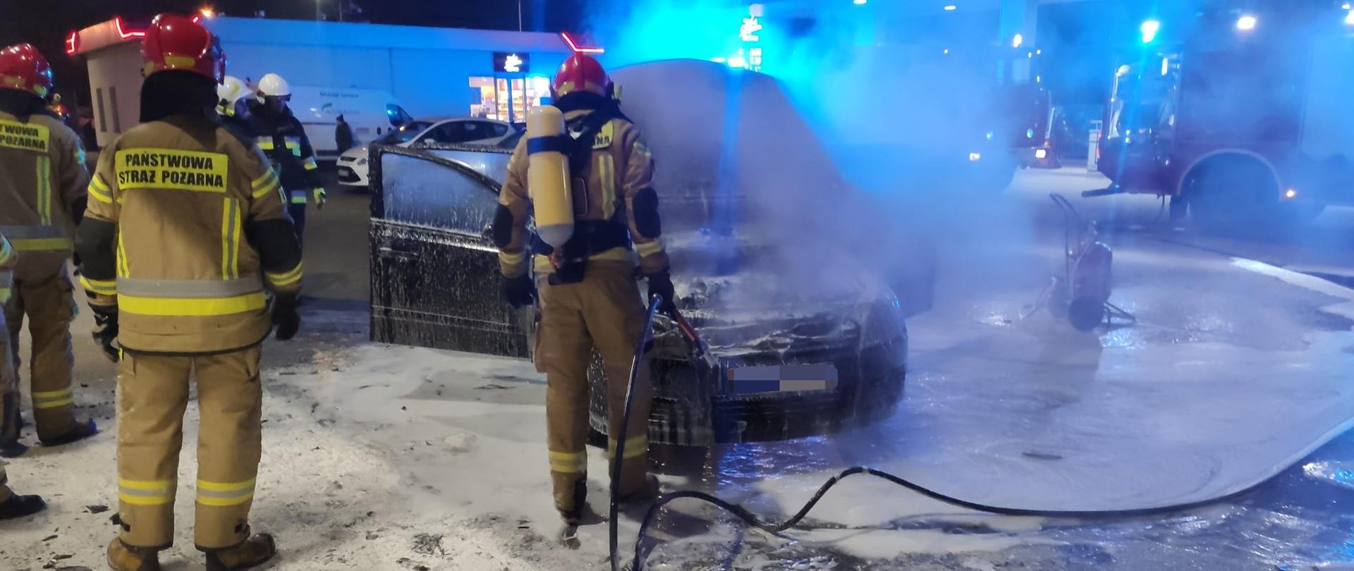 Zdjęcie przedstawia strażaków podczas akcji gaszenia pożaru samochodu osobowego. Jeden ze strażaków ma na sobie założony aparat ochrony układu oddechowego. Trzyma w ręce prądownice od linii szybkiego natarcia.