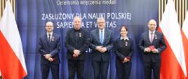 Pod wbudowanym w ścianę wielkie ekranem z napisem Ceremonia wręczenia medali „Zasłużony dla Nauki Polskiej Sapientia et Veritas” stoi czterech mężczyzn w garniturach i ubrana na czarno kobieta, po bokach polskie flagi.