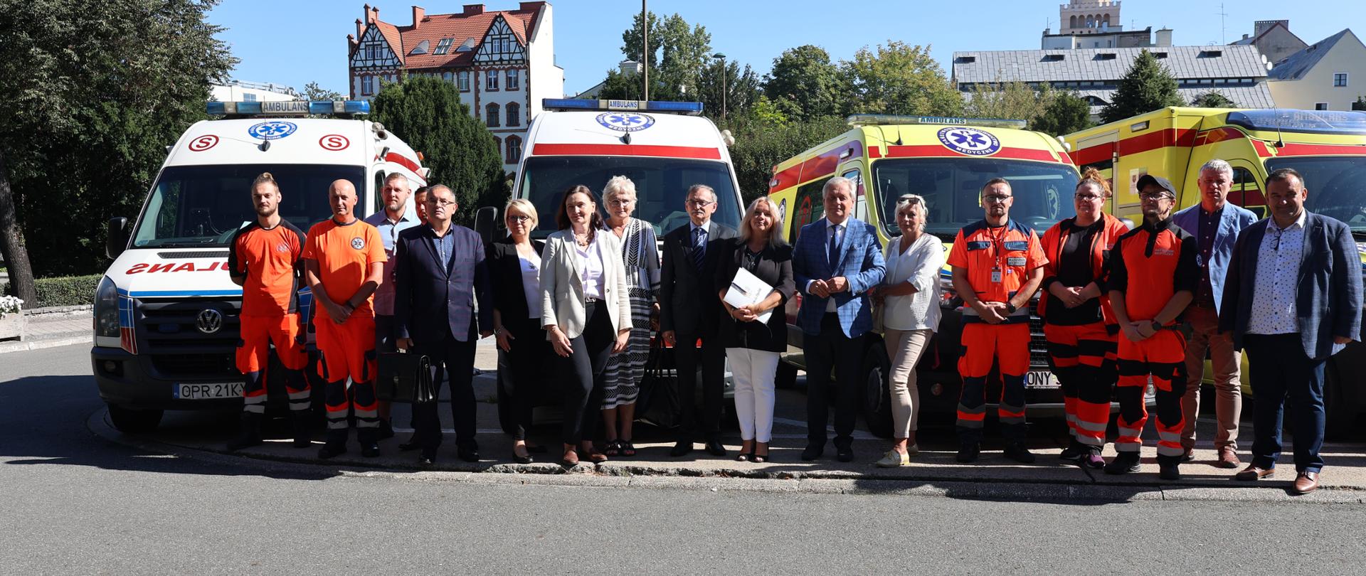 Uczestnicy dzisiejszego zdarzenia (06.09.2023 r.) stoją na parkingu przed Opolskim Urzędem Wojewódzkim w Opolu. W tle ambulansy oraz zabudowa Opola.