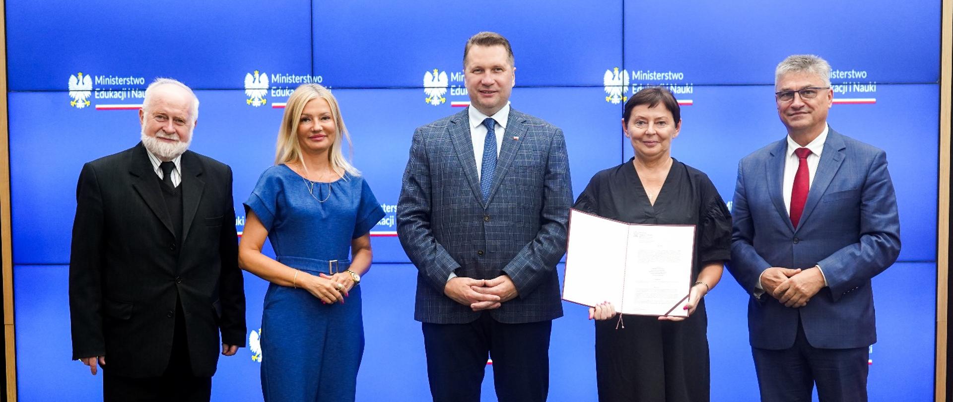 Pod ścianą stoi grupa pięciu osób, pośrodku minister Czarnek, stojąca obok niego kobieta w czarnym ubraniu trzyma otwartą teczkę z dokumentami.