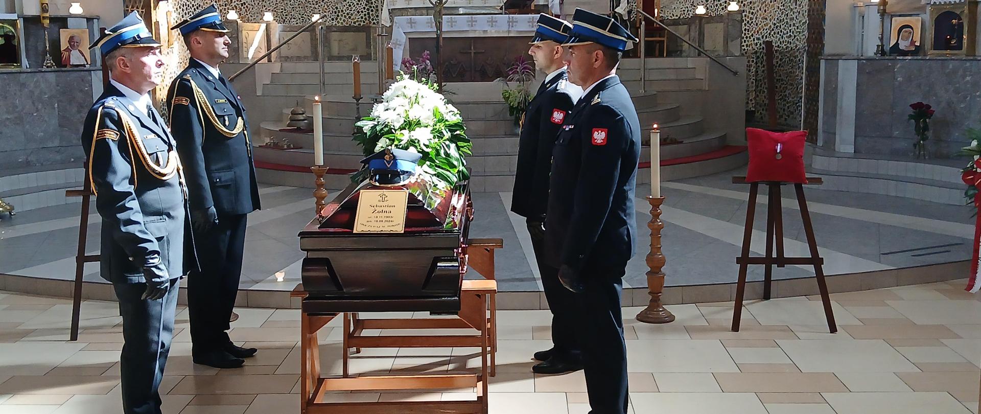 Zdjęcie przedstawia scenę wewnątrz kościoła podczas uroczystości pogrzebowej. W centrum zdjęcia znajduje się trumna przykryta wieńcem z białych kwiatów i złożonym na niej nakryciem głowy oraz odznaką. Na trumnie widoczna jest tabliczka z napisem "Ś.P. Sebastian Żołna". Czterech funkcjonariuszy Państwowej Straży Pożarnej stoi w szeregu, ustawionych na baczność przy trumnie. Są oni ubrani w pełne, formalne mundury z ciemnego materiału, z charakterystycznymi niebieskimi czapkami i złotymi akcentami, które widoczne są na mundurach. W tle widać ołtarz oraz elementy wystroju kościoła, takie jak świece i obrazy świętych. W górnej części zdjęcia, na stopniach przy ołtarzu, znajduje się ksiądz prowadzący uroczystość.
Atmosfera na zdjęciu jest poważna i pełna szacunku, co sugeruje, że jest to oficjalna uroczystość pożegnania zmarłego funkcjonariusza Państwowej Straży Pożarnej.