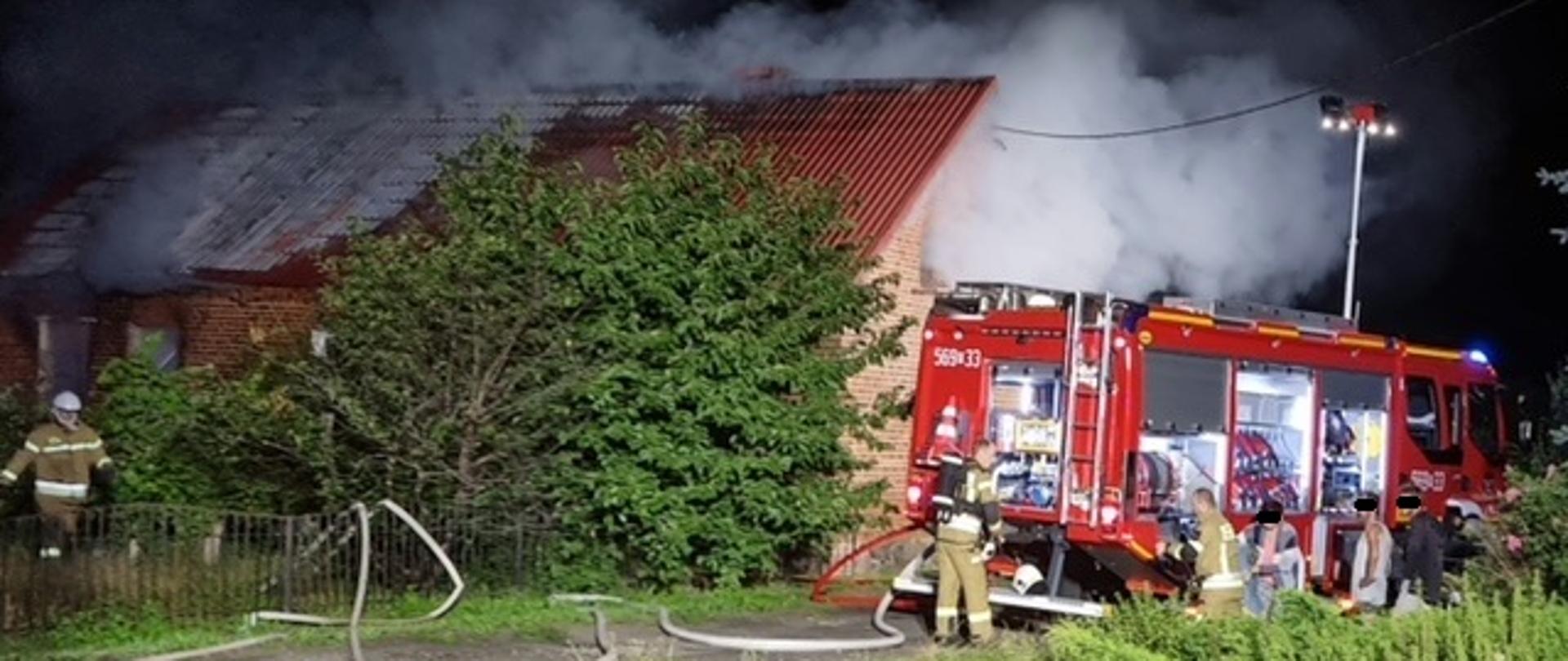 Czerwony, ceglany budynek parterowy o skośnym dachu, nad budynkiem widoczne kłęby dymu, z prawej strony znadjue się samochód gaśniczy przed którym stoją strażacy opiekujący się osobami poszkodowanymi w pożarze