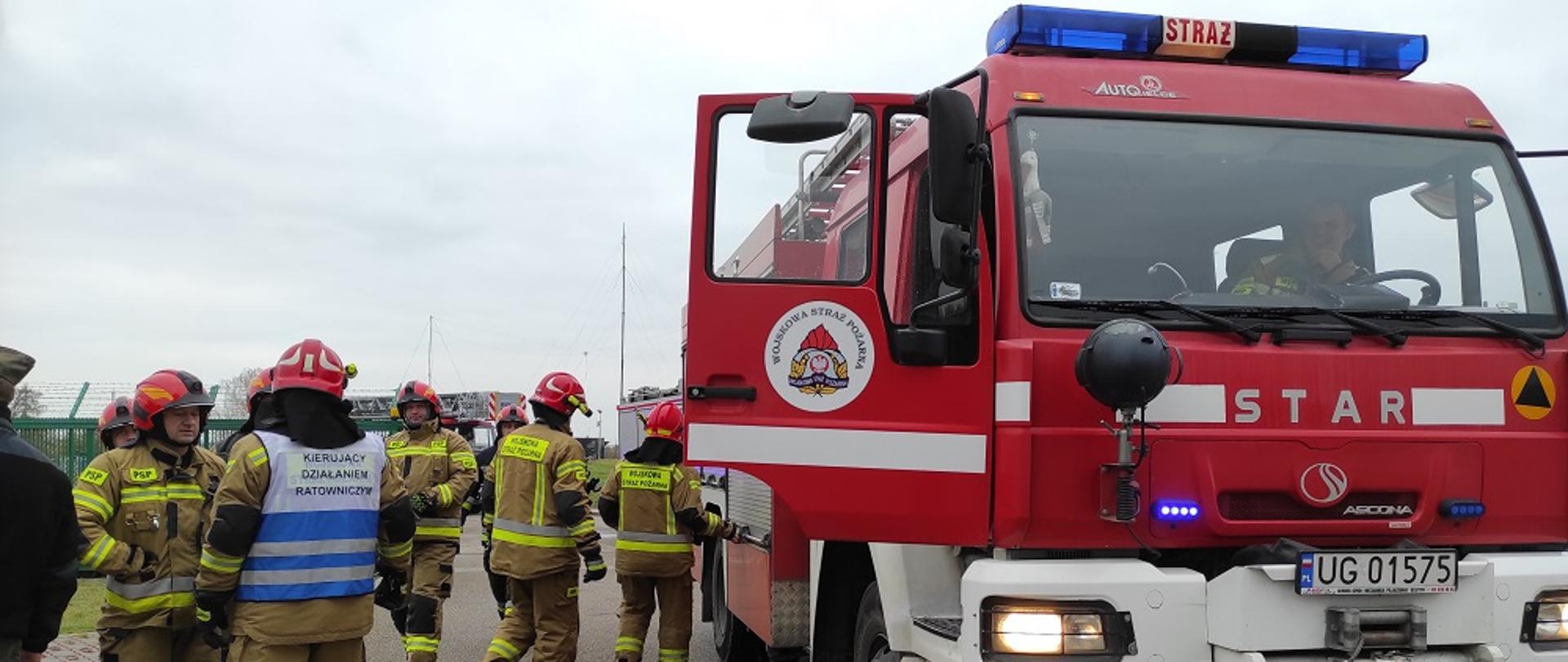 Z prawej strony czerwony samochód strażacki, z lewej strażacy przygotowują się do ćwiczeń.