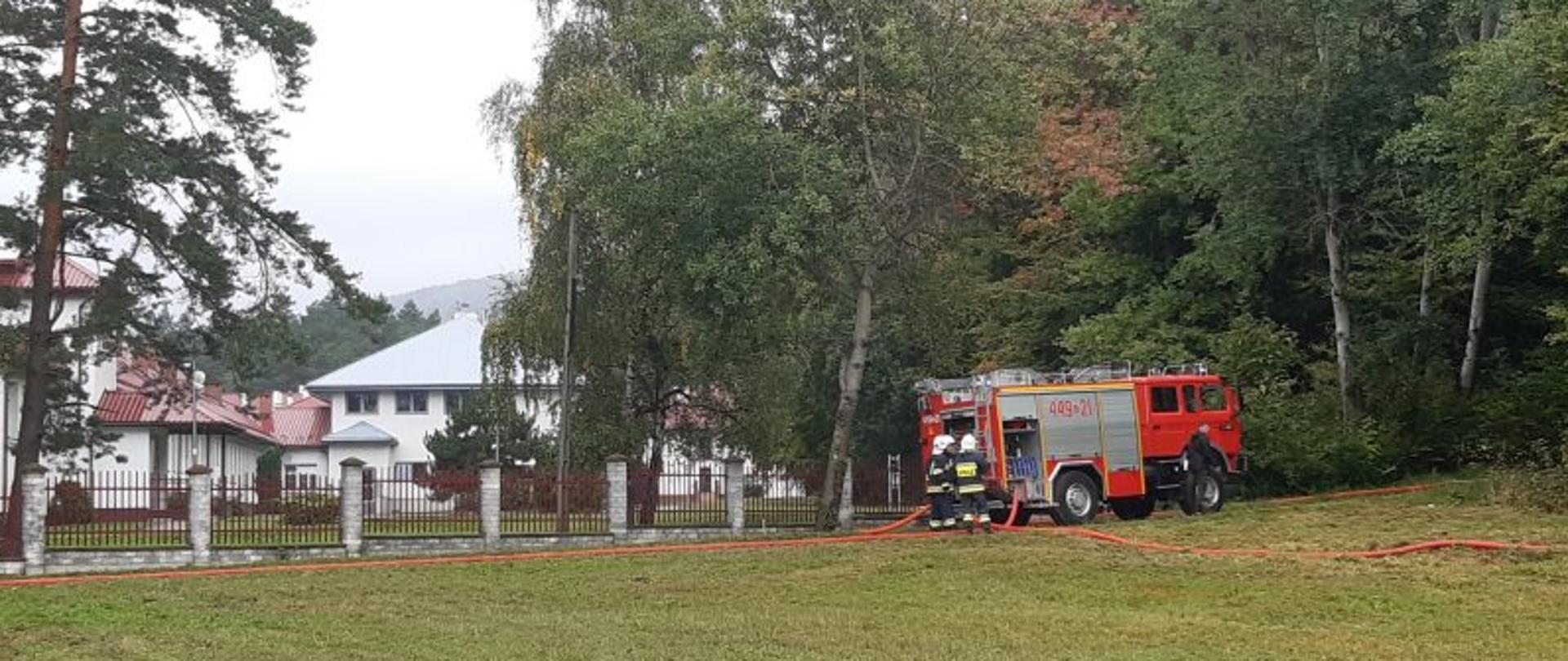 Na zdjęciu samochód ratowniczo-gaśniczy z podłączoną armaturą wodną. Przed samochodem zielona łąka. Na drugim planie za samochodem wysokie zielone drzewa oraz budynki mieszkalne
