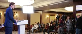 Wiceminister Jan Szyszko stoi z lewej strony przy pulpicie i mówi do mikrofonu. Z prawej strony stoją dziennikarze i kamery telewizyjne. 