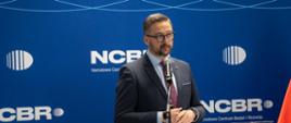 Na tle niebieskiej ścianki z napisem NCBR za przezroczystą mównicą stoi minister Kulasek i mówi do mikrofonu na stojaku.