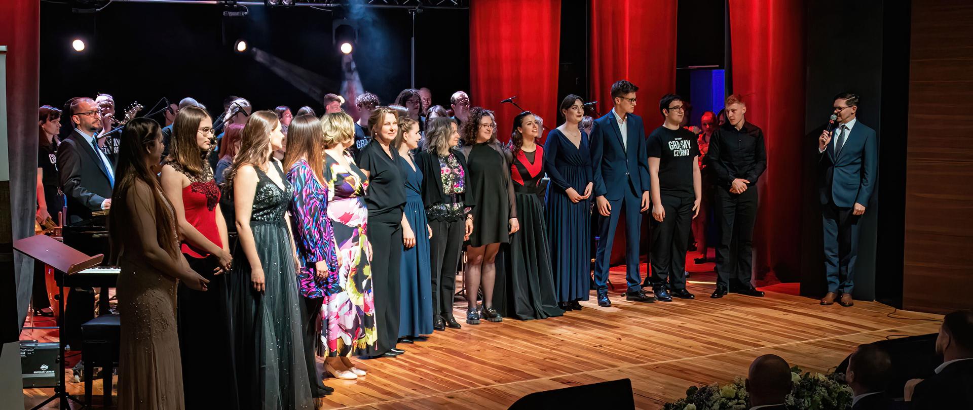 Jubileusz 50-lecia Państwowej Szkoły Muzycznej I st. im. I. J. Paderewskiego w Głubczycach. Wykonawcy koncertu zgromadzeni na scenie MOK w Głubczycach. Z prawej strony zdjęcia stoi pan dyrektor Adam Kopyto, w prawy dolnym rogu widać sylwetki osób na publiczności.