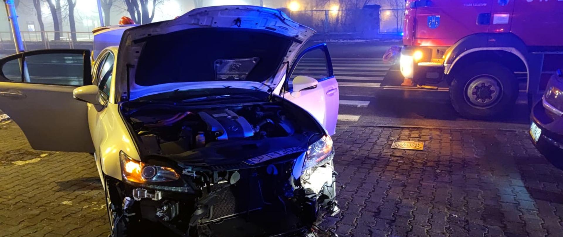 Zdjęcie przedstawia na pierwszym planie biały samochód marki Lexus z rozbitym przodem i podniesioną klapą silnika. Obok stoi średni samochód ratowniczo - gaśniczy. Za samochodem Lexus widać przejście dla pieszych.