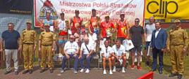 strażacy oraz organizatorzy zawodów, na podium stoją zwycięzcy zawodów za nimi na siatce banery reklamowe