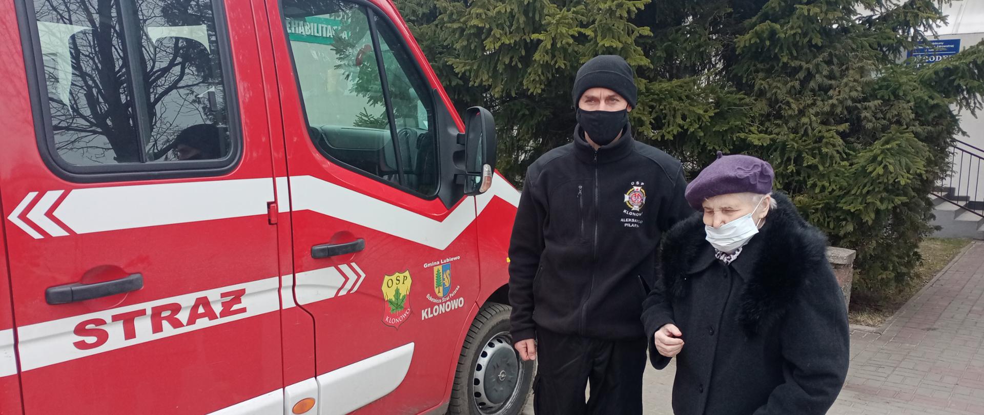 Samochód strażacki jednostki Ochotniczej Straży Pożarnej w Klonowie, obok druh z Ochotniczej Straży Pożarnej w Klonowie i pacjentka przywieziona na szczepienie przeciwko COVID-19.
W tle budynek Ośrodka Zdrowia w Bysławiu.