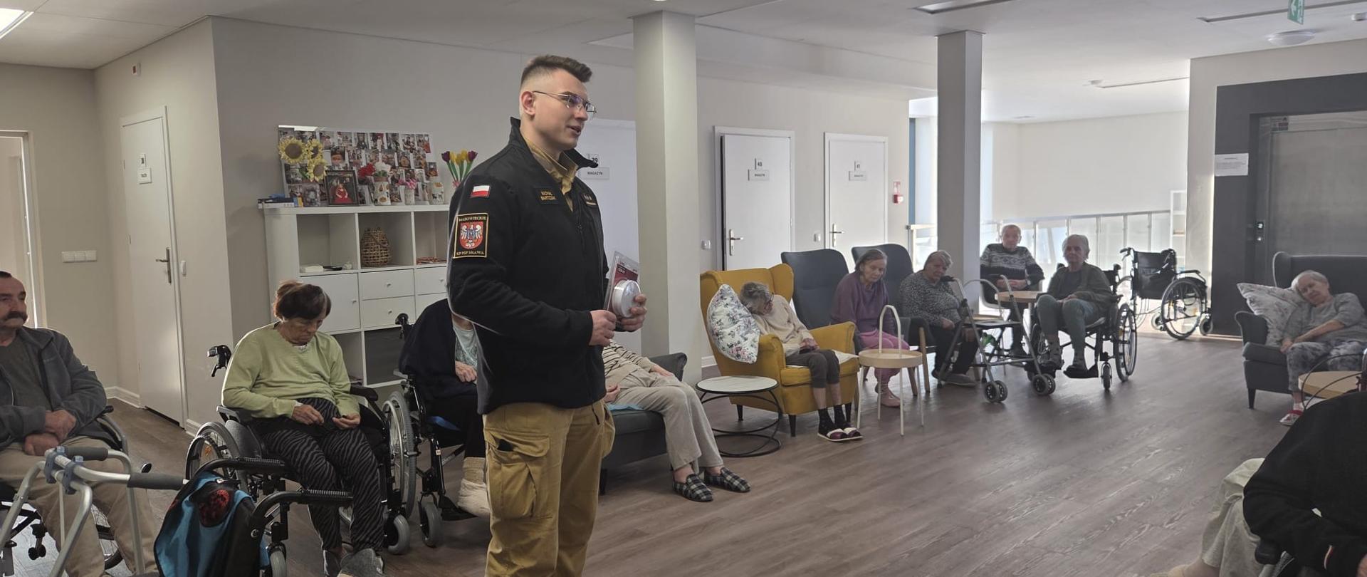 W jasnej sali domu opieki stoi strażak w służbowym umundurowaniu. W dłoniach trzyma czujkę dymu i prowadzi spotkanie edukacyjne. Wokół niego siedzą starsze osoby, część na wózkach inwalidzkich, uważnie słuchając jego wypowiedzi. W tle znajdują się regały z dekoracjami oraz drzwi do innych pomieszczeń.