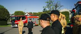 Uroczysta zbiórka z okazji podniesienia flagi, pododdział strażaków stoi na tle samochodów pożarniczych, komendant przemawia do strażaków