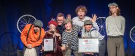 Oświetlona na niebiesko scena teatralna, a na niej trzy dziewczyny w makijażu i strojach scenicznych w rękach trzymają dwa oprawione dyplomy. Za nimi i wokół nich są dwaj chłopcy i dwaj mężczyźni. Wszyscy pozują do zdjęcia pochylając się do przodu i robiąc "głupie miny".