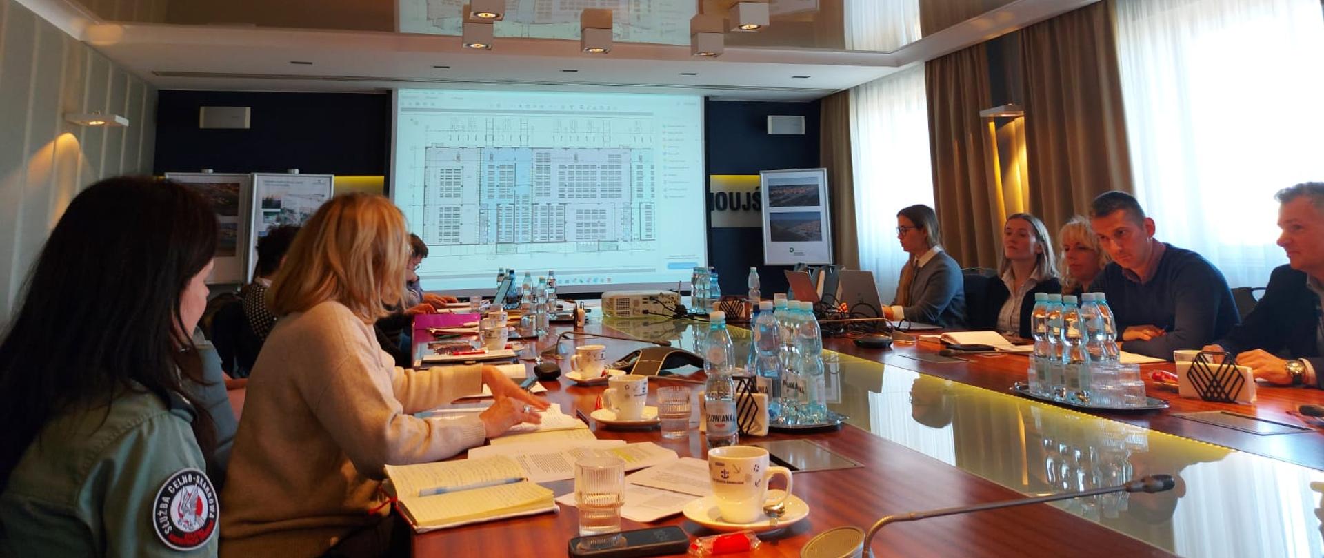 Zdjęcie przedstawia salę konferencyjną z dużym, prostokątnym stołem pośrodku. Po obu stronach stołu usadowione są osoby biorące udział w konferencji. Na ścianie sali konferencyjnej wyświetlany jest pokaz slajdów.