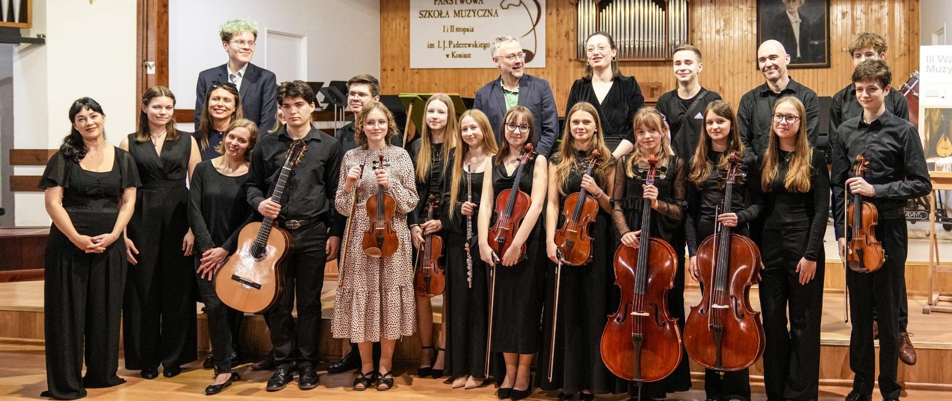 Pamiątkowe zdjęcie grupowe uczestników i wykładowców III Warsztatów Muzyki Dawnej w Koninie, wykonane na scenie auli szkolnej z instrumentami takimi jak skrzypce, wiolonczele, flet i gitara barokowa, na tle organów i tablicy z nazwą szkoły.