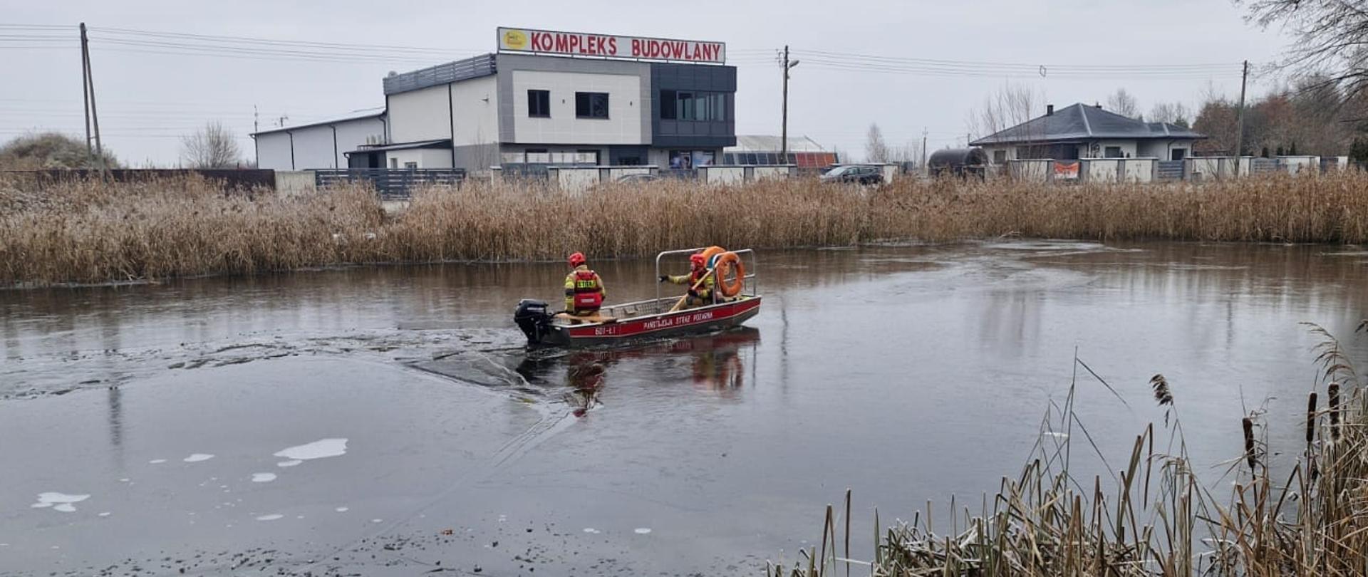 Na zdjęciu widoczna jest łódź ratownicza z dwoma strażakami - ratownikami na pokładzie. Płynie po częściowo zamarzniętym akwenie wodnym, otoczonym suchymi trzcinami. W tle widoczny jest kompleks budowlany oraz inne zabudowania. Strażacy z Jednostki Ratowniczo - Gaśniczej Państwowej Straży Pożarnej w Przysusze przy użyciu łodzi ratowniczej podpływają do przymarzniętego łabędzia i go uwalniają. 
