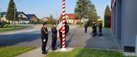 Trzech strażaków stoi obok masztu flagowego i jeden ją przypina. Pozostali strażacy stoją w dwuszeregu. W tle zdjęcia budynek garażu Komendy z napisem Państwowa Straż Pożarna.