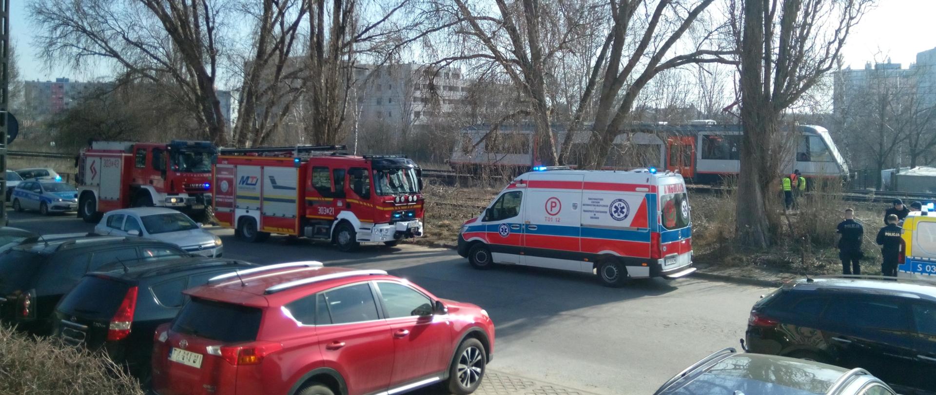 Zdjęcie przedstawia pojazdy służb ratowniczych zaparkowane na ulicy Mielczarskiego, a w głębi na torowisku stoi pociąg osobowy.