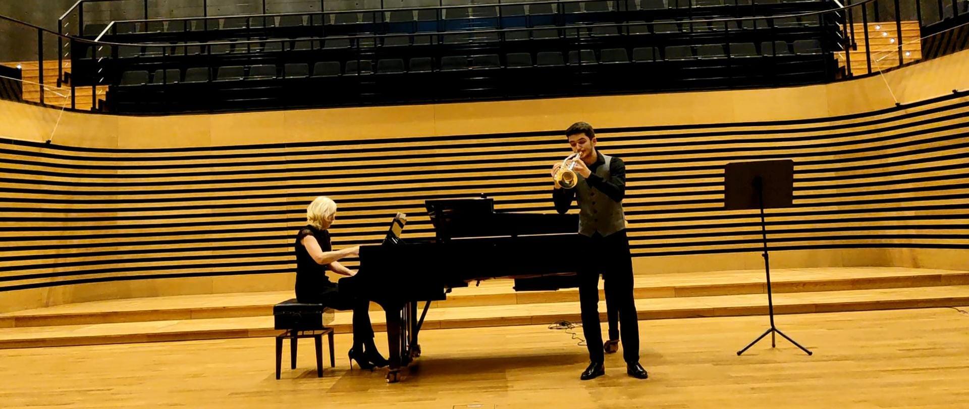 Zdjęcie dużej sceny koncertowej o żółtawej kolorystyce (parkiet i okalające ją wyciszenie). Na scenie młody, ubrany na czarno i w szarą kamizelkę trębacz i akompaniatorka ubrana na czarno o blond włosach towarzysząca mu na fortepianie.