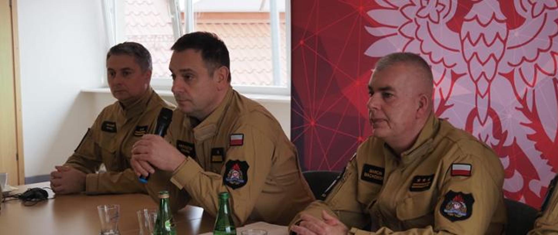 2 komendantów wojewódzkich i naczelnik wydziału siedzą przy stole. Komendant trzyma w ręce mikrofon .W tle widać orła. 